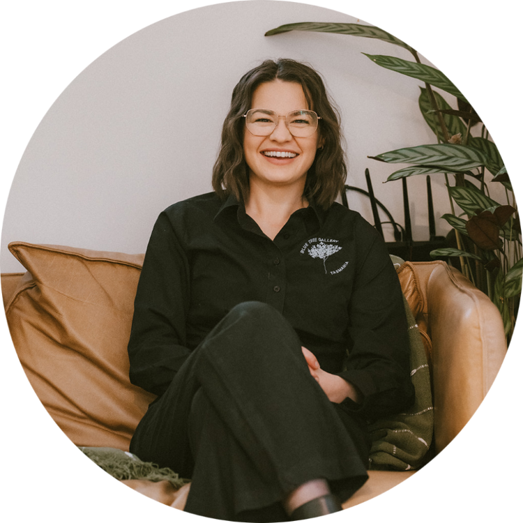 Artist Aimee Rowland - A woman with short brown hair and glasses, wearing a black shirt with a logo and black pants, sitting in her shipping container art gallery on a brown leather chair