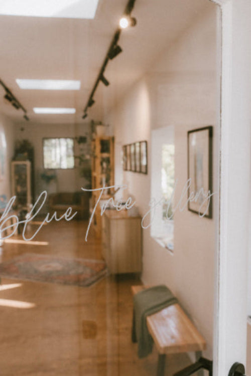 The view of the inside of Blue Tree Gallery through the glass door in focus is the white writing on the door which reads "blue tree gallery" in cursive