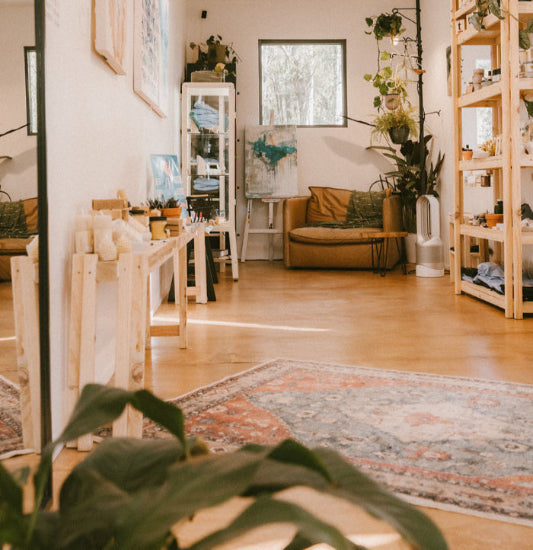 The inside of blue Tree Gallery (White walls and timber floor) with timber shelving on both sides, a rug on the floor and in focus at the end of the long thin gallery is a brown leather chair, plants and an abstract painting.