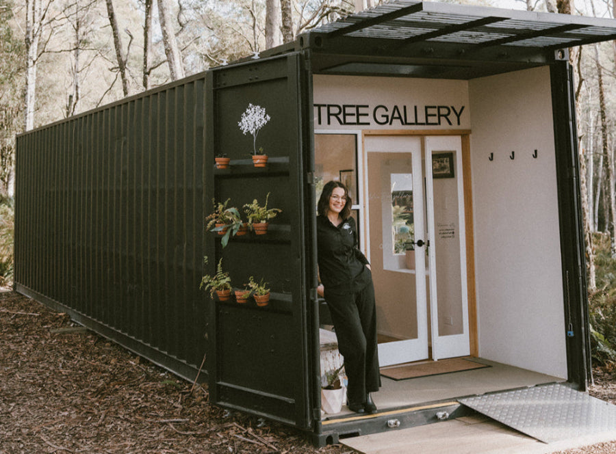 The artist and owner of Blue Tree Gallery Aimee Rowland dressed in black standing outside her Renovated Shipping Container Art Gallerysurrounded by Tasmanian wilderness with potted plants on the door, and painted black with a ramp leading to the entrance of the art gallery