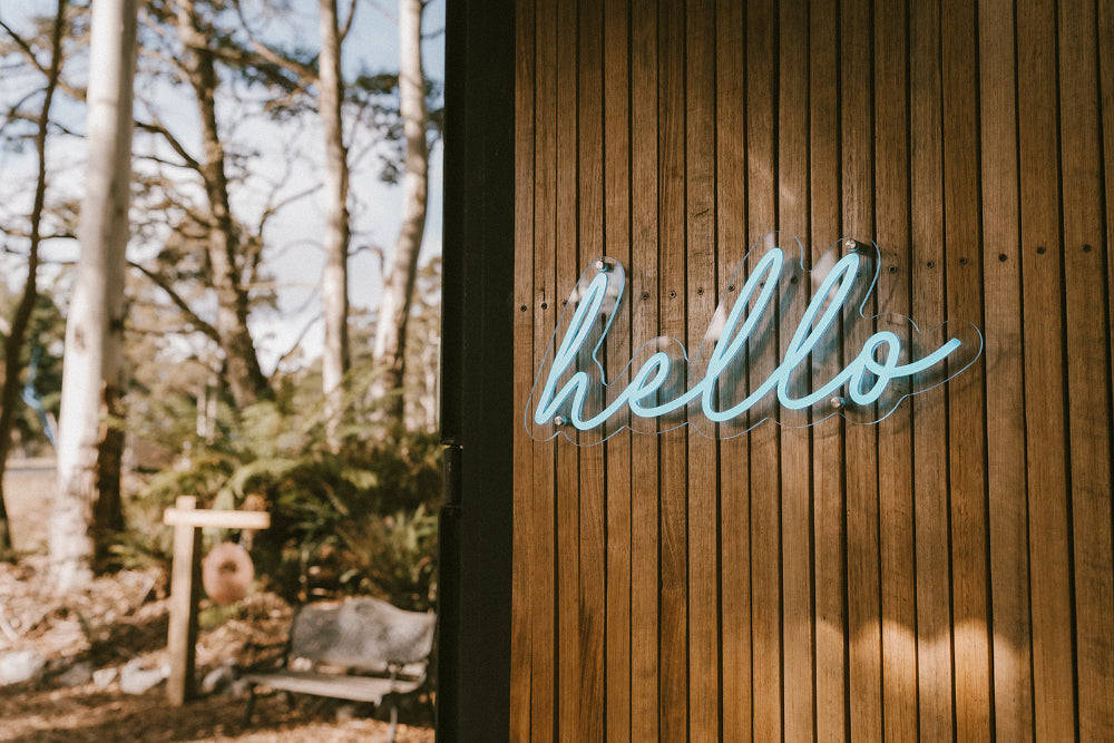 Timber slated wall with a neon blue sign saying "hello" in cursive, in the background to the left are the trees ferns and a seat out of focus 